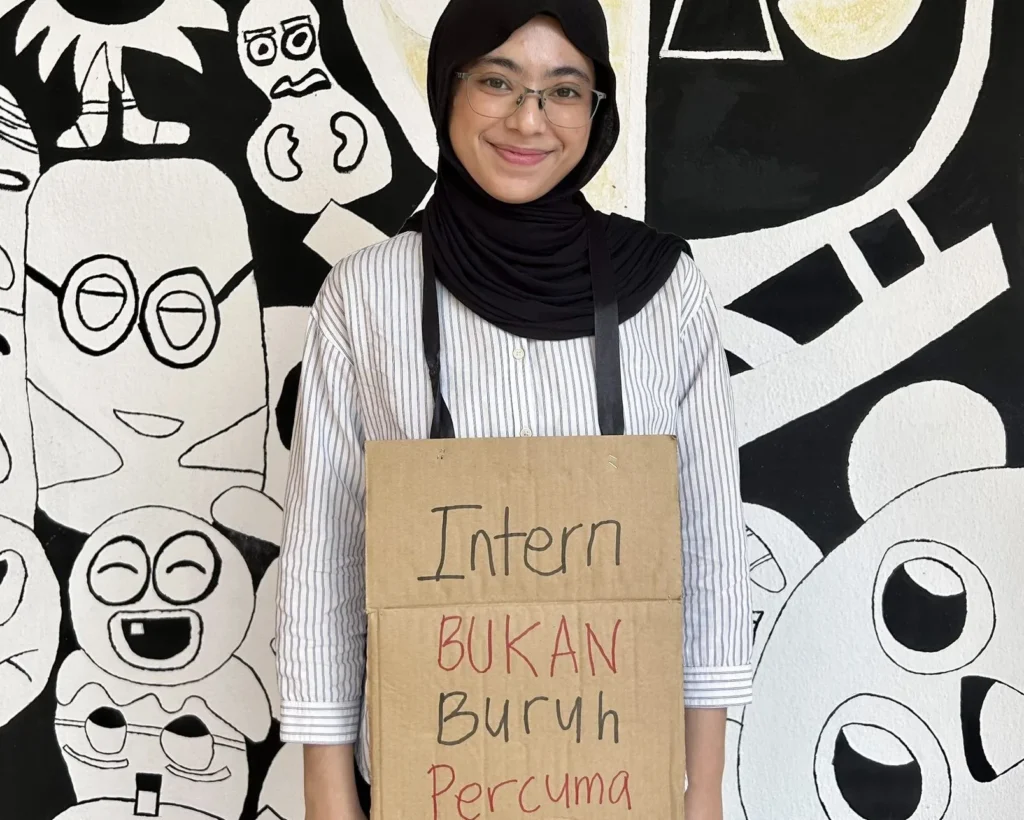 Aktivis bergambar dengan sepanduk bertulis "Pelatih Industri (Intern) Bukan Buruh Bercuma".